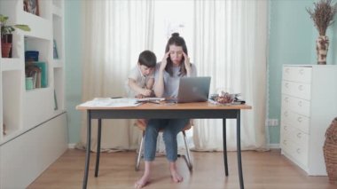 Ebeveynler evde çalışıyor. Uzaktan kumanda. Aile bireysel izolasyonda. Merkez ofis. Masada bir kadın çalışıyor. Oğlu annelerin işine karışıyor. Annem yorgun. Karantinaya alın. Salgın hastalık. Kriz.