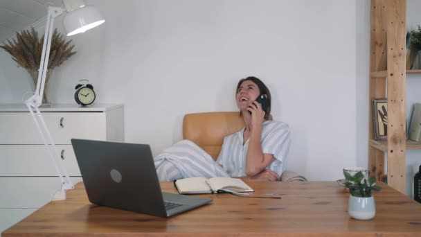 女性がノートパソコンの椅子に座って電話で話している。パジャマ姿の若い女性。フリーランス。自己分離。ビジネスレディー。家で働いてる。オンライン相談家庭教育 — ストック動画