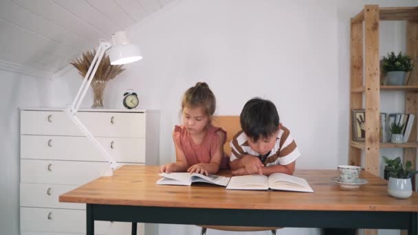 Ein Junge und ein Mädchen lesen ein Buch, das auf einem Tisch liegt. Die Kinder machen ihre Hausaufgaben. Bildung zu Hause. Selbstisolierung. Kinderstunden. Zeitlupe — Stockvideo