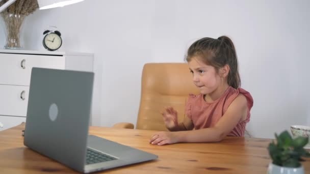 A rapariga olha para o portátil. A criança se comunica online enquanto está sentada à mesa. A criança aprende online. Educação doméstica. A luz de uma lâmpada de mesa. Treinamento online Auto-isolamento Movimento lento — Vídeo de Stock