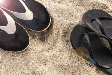 Erkekler ve kadınların plaj ayakkabıları güneşin altında, kumun üzerinde. İki çift siyah parmak arası terlik.