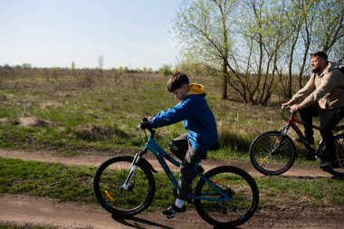 Baba ve küçük oğul çayırda bisiklete biniyorlar.