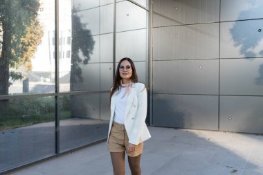 Gri, modern bir binanın arka planında beyaz ceketli, gözlüklü, kendine güvenen bir iş kadını.