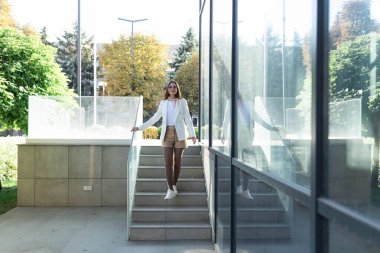 Gözlüklü, beyaz ceketli, beton merdivenli, kendine güvenen bir iş kadını.