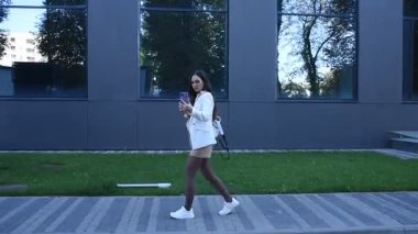 Beyaz ceketli ve sırtında sırt çantasıyla genç bir kadın modern bir binanın önünden geçerken görüntülü konuşma yapıyor.