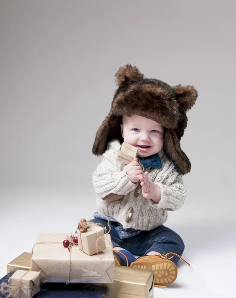 Bebé divertido en un sombrero de piel de invierno con cajas de regalo sobre backgro gris —  Fotos de Stock