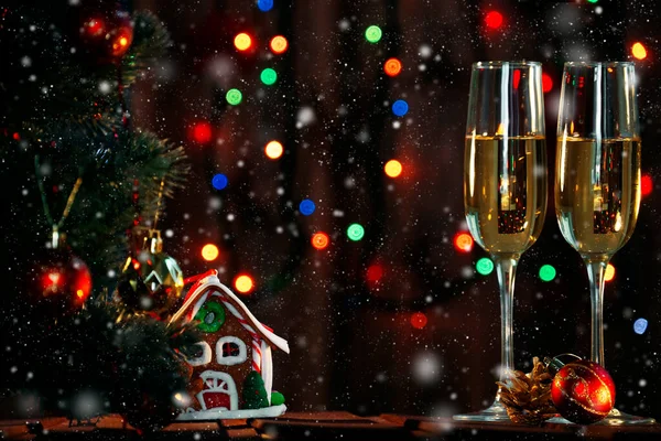 Noel ağacı, oyuncak zencefilli kek evi ve üzerinde siyah bir çelenk olan masanın üstünde Noel süsleri olan iki şampanya bardağı.
