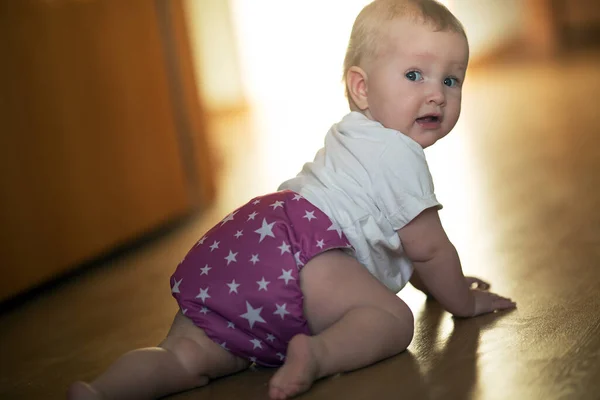 Menina bebê vestindo fraldas reutilizáveis. Menina está rastejando no chão sob a luz do sol — Fotografia de Stock