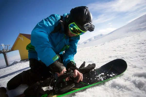 Snowboarder in bright sportive clothes repairing his snowboard on the mountain top — Stock Photo, Image