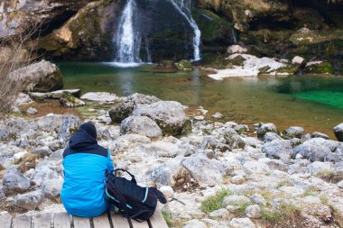 Parlak mavi spor ceketli genç adam şelalenin yanında dinleniyor. İlkbaharın başlarında, Slovenya. Ruh sağlığı konsepti