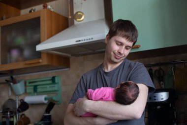 Genç bir adam yeni doğmuş kızını dikkatli bir şekilde tutuyor. Adam mutfakta duruyor ve gülümsüyor.