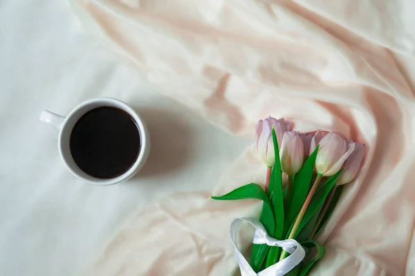 Bouquet of tulips and cup of coffee lies on the background of silk fabric with folds.