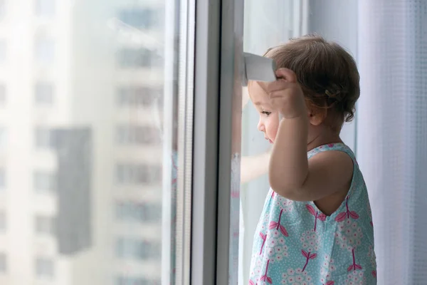 Pequeña chica pensativa contra la ventana cerrada mirando hacia fuera. Concepto de cuarentena y autoaislamiento. Copiar espacio — Foto de Stock