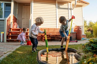 Bahçede oynayan çocuklar su pompası ile küçük bir göletin yanında.