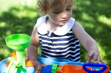 Cute toddler girl playing with water toy outdoors. Summer sunny day, baby is wearing striped dress