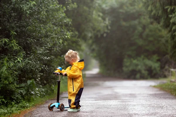 Little blonde girl wearing yellow waterproof overall standing outdoors in countryside with scooter — Photo