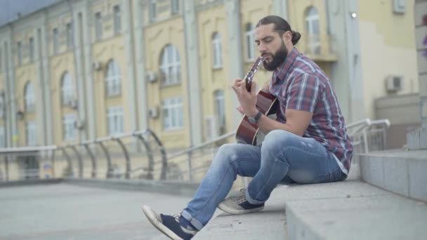 Wide shot side view of young Caucasian man sitting on urban stairs and playing guitar. Handsome bearded musician performing on city street outdoors. Lifestyle concept. — Stock Video