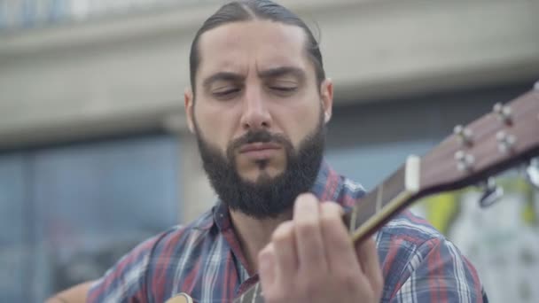 Närbild porträtt av fängslad kaukasisk musiker som spelar gitarr utomhus. Snygg absorberad man som uppträder i urban stad. Hobby och livsstil. — Stockvideo