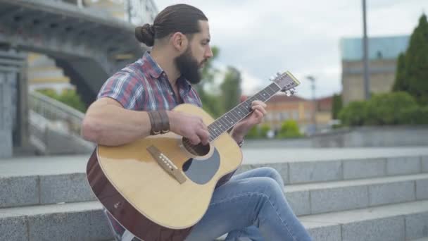 Widok z boku portret młodego człowieka grającego na gitarze siedzącego na miejskich schodach. Skoncentrowany kaukaski gitarzysta występujący samotnie w letni dzień na świeżym powietrzu. Koncepcja sztuki i stylu życia. — Wideo stockowe