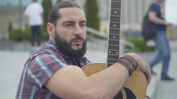 Handsome Caucasian guitarist sitting on urban street and looking away. Portrait of confident young thoughtful man with guitar outdoors. Creative guy in city. — Stock Video