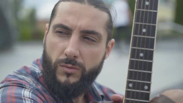 Headshot of handsome musician with guitar fingerboard. Close-up portrait of thoughtful Caucasian bearded brunette man sitting outdoors in urban city and thinking. — Stock Video