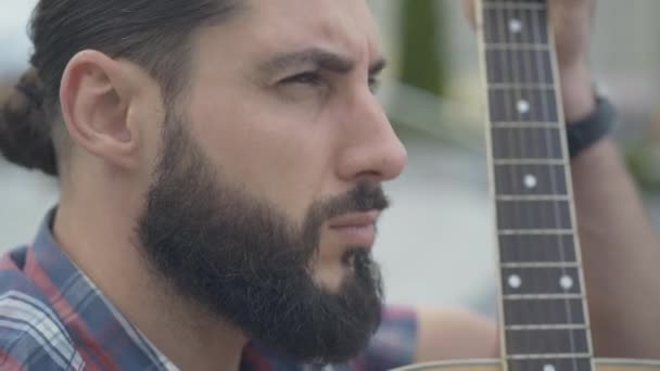 Close-up of bearded engrossed guitarist playing outdoors and shaking head. Side view portrait of handsome absorbed Caucasian man playing musical instrument in city. — Stock Video