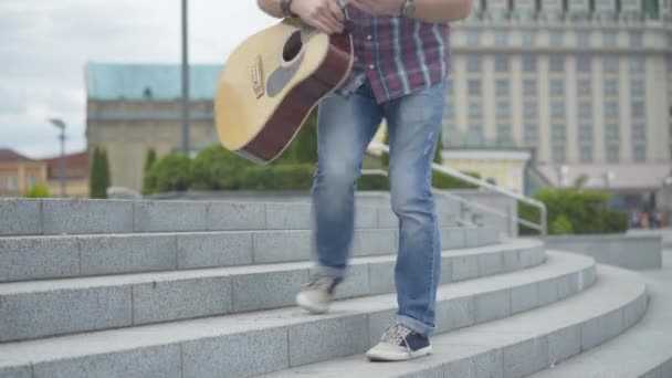 Caucasian man walking up urban stairs, taking off hat, and start playing guitar. Portrait of street musician performing to earn money outdoors on summer day. Begging concept. — Stock Video