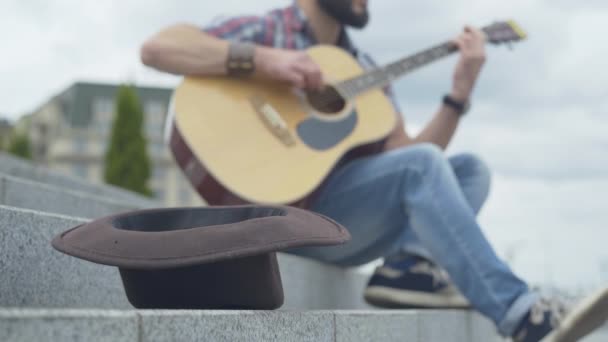 Zbliżenie brązowego kapelusza na miejskich schodach z rozmazanym człowiekiem grającym na gitarze w tle. Nierozpoznany kaukaski gitarzysta występujący w mieście w letni dzień. — Wideo stockowe