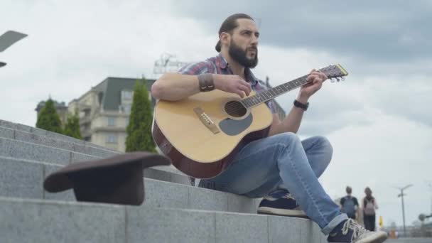 Widok z boku męskiego muzyka występującego w miejskim mieście w pochmurny letni dzień. Portret skoncentrowanego białego mężczyzny grającego na gitarze siedzącego na schodach na zewnątrz. — Wideo stockowe