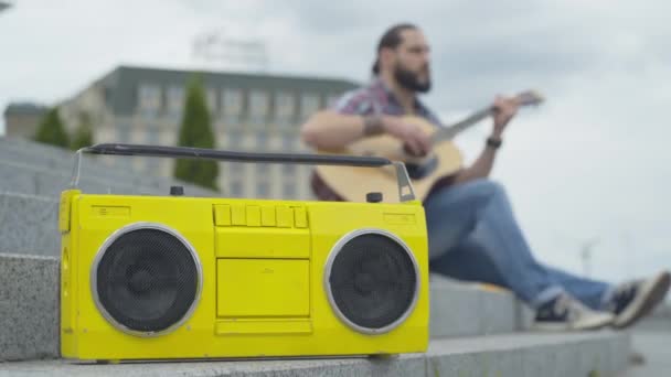 Vintage yellow tape recorder standing on urban stairs with blurred guitarist playing at the background. Young Caucasian musician performing on city street for money. Poverty concept. — Stock Video