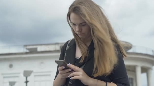Konzentrierte Kaukasierin, die draußen auf dem Smartphone Nachrichten tippt. Porträt der jungen schönen brünetten Dame, die an windigen Tagen auf der Straße steht und online Nachrichten verschickt. — Stockvideo
