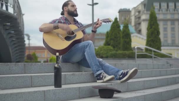 Brett fotograferat porträtt av entusiastisk musiker som spelar gitarr på stadstrappor med vinflaska och hatt på framsidan. Stackars kaukasier som uppträder utomhus för pengar. Fattigdom och livsstil. — Stockvideo