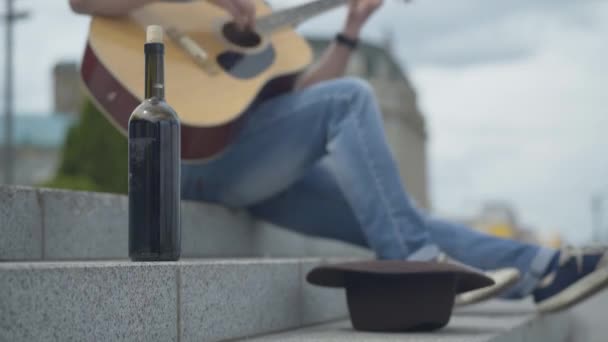 Wine bottle and beggar hat on urban stairs with blurred enthusiastic musician playing guitar at the background. Unrecognizable Caucasian man performing outdoors earning money. — Stock Video