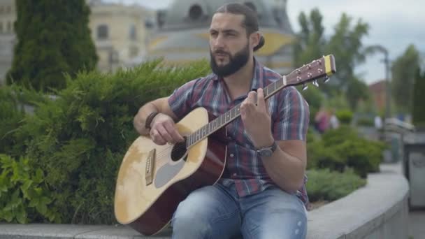 Middle shot of serious Caucasian handsome man enjoying playing guitar outdoors. Portrait of confident young male musician performing on urban city street on windy summer day. — Stock Video