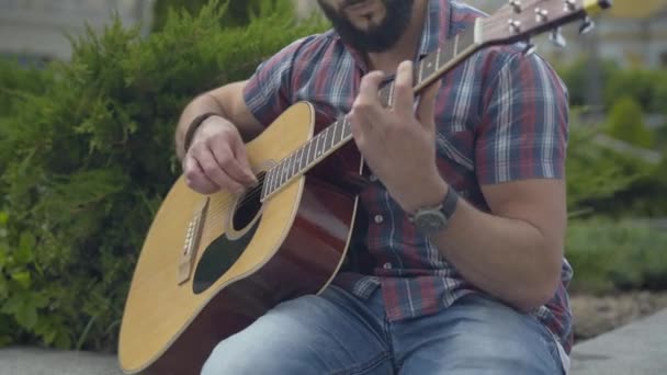 Unrecognizable bearded male musician performing on summer day. Young Caucasian man playing guitar in urban city. Guitarist enjoying hobby outdoors. — Stock Video