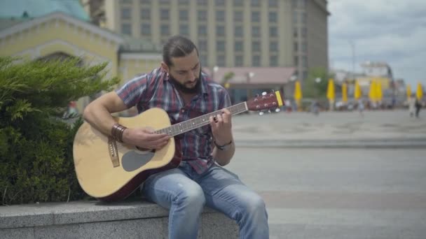 Koncentrerad gitarrist spelar musikinstrument utomhus med suddig urban stad i bakgrunden. Porträtt av fokuserad ung kaukasisk man njuter av musik. — Stockvideo