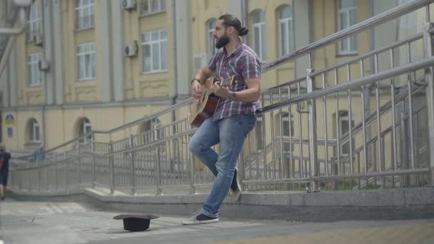 Wide shot of poor Caucasian man standing on city street and playing guitar. Portrait of unhappy musician begging money for living. Beggary and poverty in modern society. — Stock Video