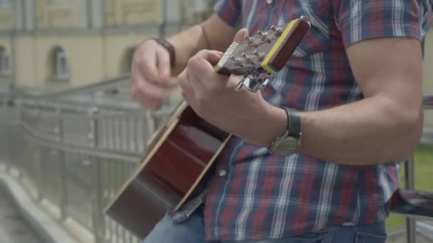 Unrecognizable man standing at urban fence and playing guitar. Hands of male musician playing stringed musical instrument outdoors on summer day. — Stock Video