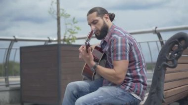 Yaz günü kentteki bankta oturan konsantre gitaristin yan görüntüsü. Açık havada müzik aleti çalan düşünceli beyaz bir adamın portresi..