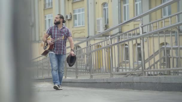 Tankeväckande kaukasisk man går längs den europeiska stadsgatan med gitarr och hatt. Porträtt av sorgsen manlig tiggare som strosar utomhus på sommardagen. Fattigdom och tiggeri. — Stockvideo