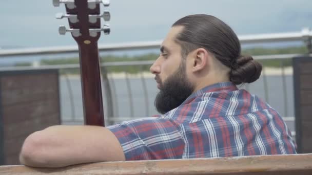 Back view portrait of thoughtful young bearded man sitting on bench with guitar and looking away. Upset Caucasian musician resting on city embankment on summer day. — Stock Video