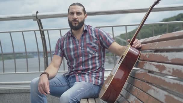 Tankeväckande självsäker man sitter med gitarr på bänken och tittar bort. Porträtt av ung vacker skäggig vit gitarrist tänker utomhus på molnig sommardag. — Stockvideo