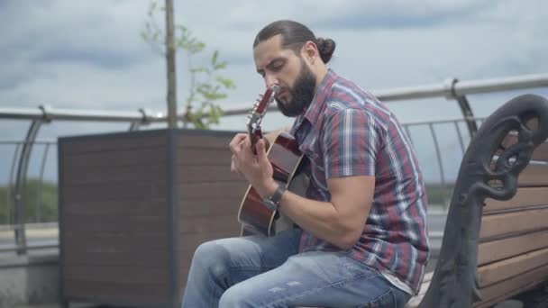 Sidovy över koncentrerad gitarrist sitter på urban bänk på sommardagen. Porträtt av omtänksam ung vit man som spelar musikinstrument utomhus. — Stockvideo