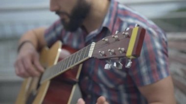 Bulanık sakallı adamın müzik aleti çaldığı gitar klavyesinin yakın çekimi. Tanınmayan beyaz müzisyen gitarist bulutlu bir yaz gününde şehir bankında oturuyor..