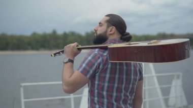 Omuzlarında gitarla nehrin yanında temiz havanın tadını çıkaran kendine güvenen beyaz bir adamın arka görüntüsü. Mutlu rahat müzisyen dışarıda dikilip başka tarafa bakıyor. Yalnızlık ve sanat konsepti.