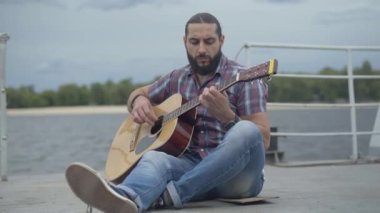 Yetenekli gitaristin portresi set seti üzerinde oturuyor ve telli enstrüman çalıyor. Şehir nehir kıyısında gitar çalan düşünceli, yakışıklı beyaz bir adam..