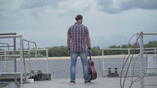 Back view of carefree musician standing on embankment and stretching hands. Wide shot of happy relaxed Caucasian man with guitar enjoying windy summer day outdoors. Happiness and lifestyle. — Stock Video