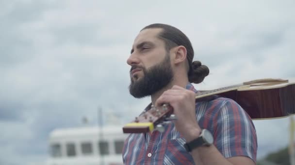 Close-up of bearded handsome man with guitar looking away and thinking. Young Caucasian musician standing with stringed musical instrument outdoors with cloudy sky at the background. — Stock Video