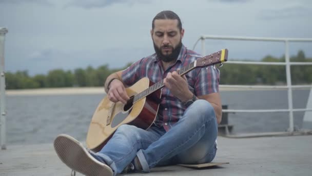 Porträt eines talentierten Gitarristen, der auf einem Damm sitzt und Saiteninstrument spielt. Nachdenklicher gutaussehender kaukasischer Mann spielt draußen am städtischen Flussufer Gitarre. — Stockvideo