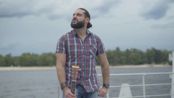 Porträt eines gutaussehenden kaukasischen Musikers, der mit Gitarre auf einem städtischen Damm steht und sich umschaut. Nachdenklicher junger Mann im Hintergrund des sommerlichen Flusses an einem windigen Tag. — Stockvideo
